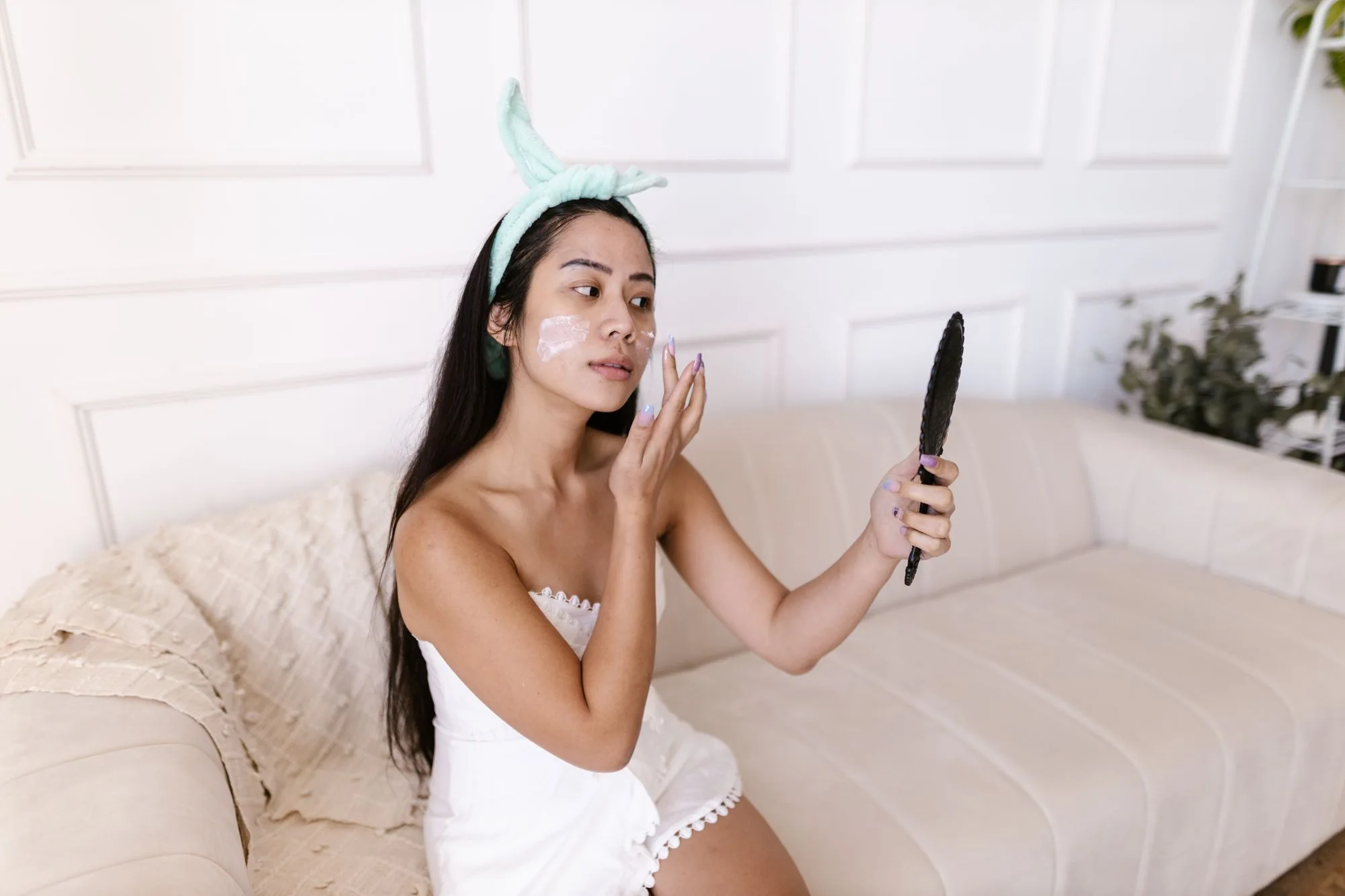 Asian woman in a towel applying face cream while looking in a handheld mirror indoors. Editorial photograph for our skincare before makeup guide for Dubai shoppers.