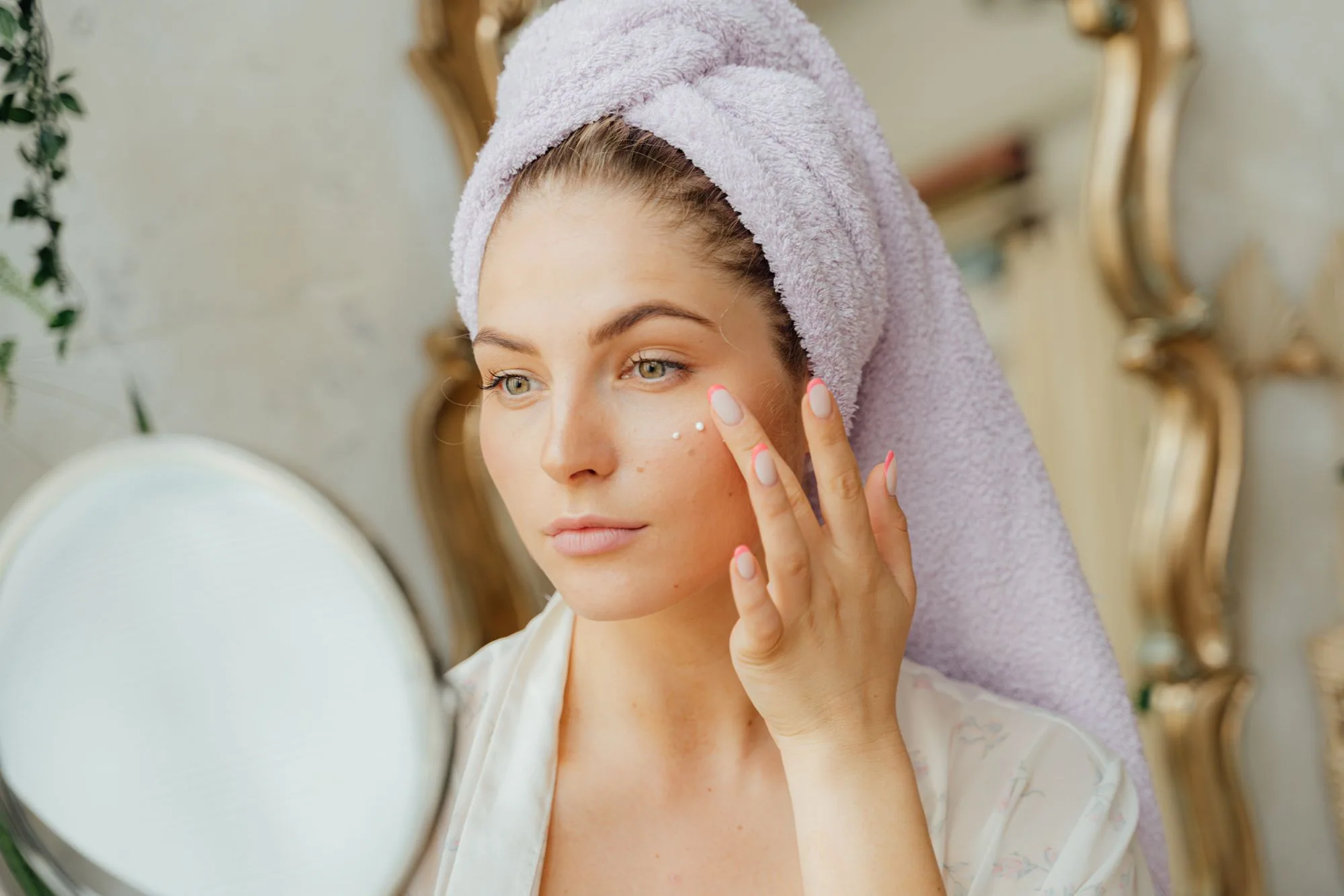 Woman with head towel applying moisturizer in front of mirror indoors. Editorial photograph for our short routine guide for Dubai shoppers.