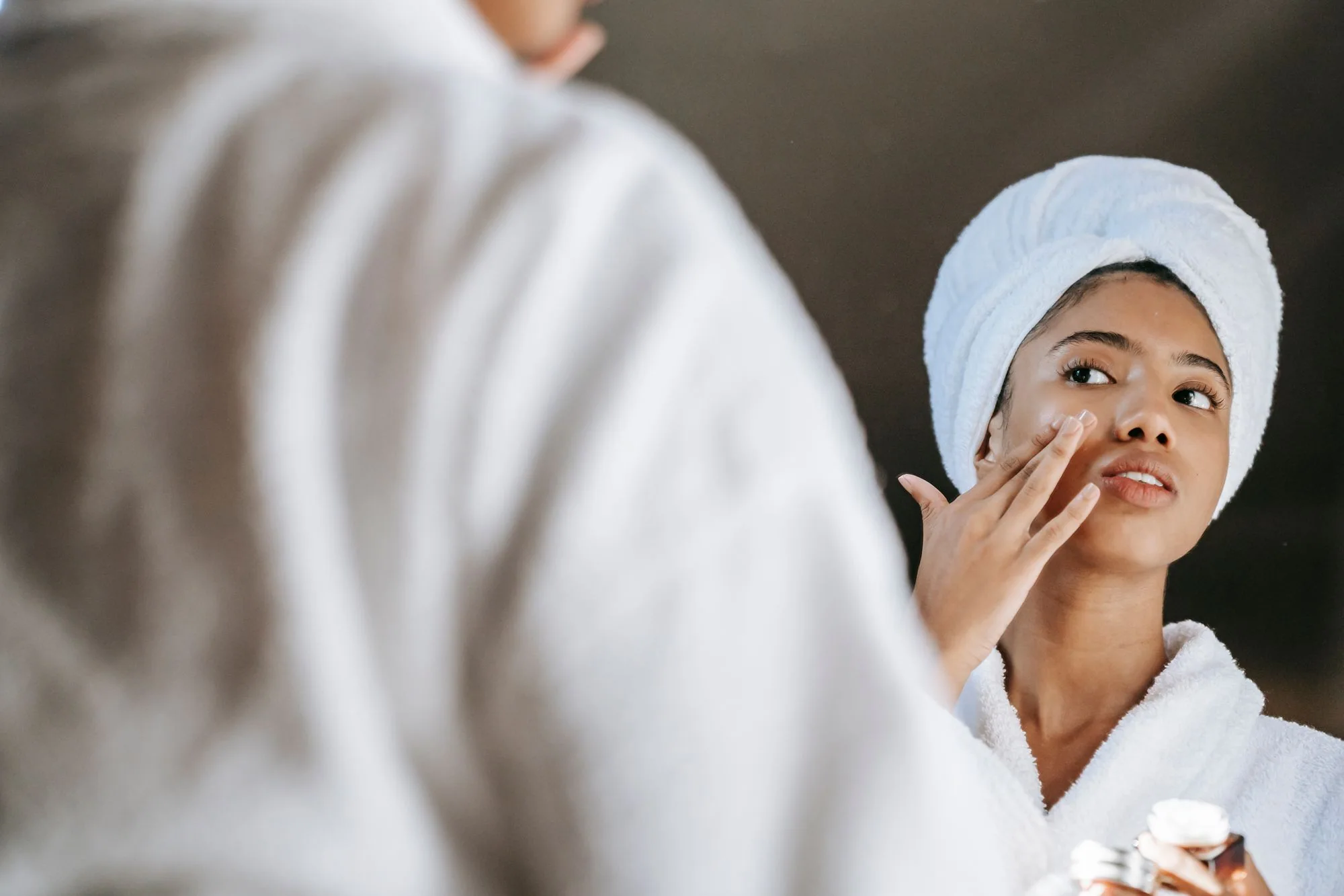 A young woman applies skincare cream while wearing a bathrobe in front of a mirror. Editorial photograph for our Moisturizing Lotion guide for Dubai shoppers.