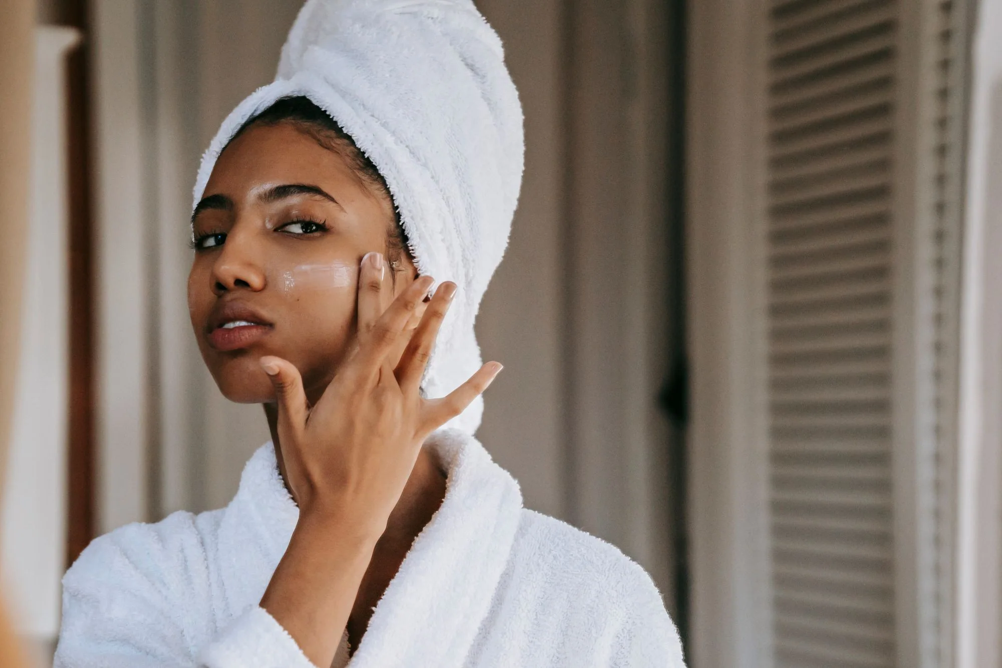 A young woman in a bathrobe applies moisturizer while looking at herself in the mirror at home. Editorial photograph for our sunscreen and makeup guide for Dubai shoppers.