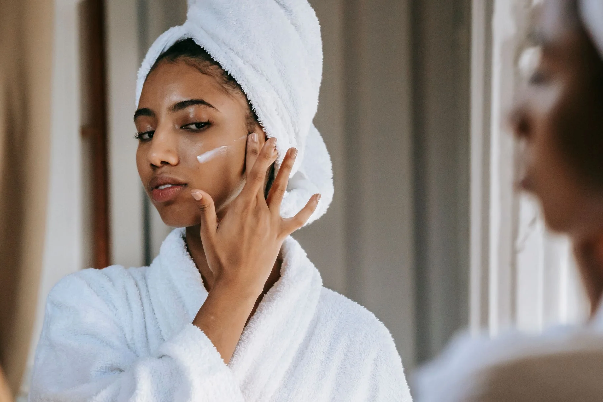 Young woman in bathrobe applying moisturizing cream while looking in the mirror at home. Editorial photograph for our Rich Cream guide for Dubai shoppers.