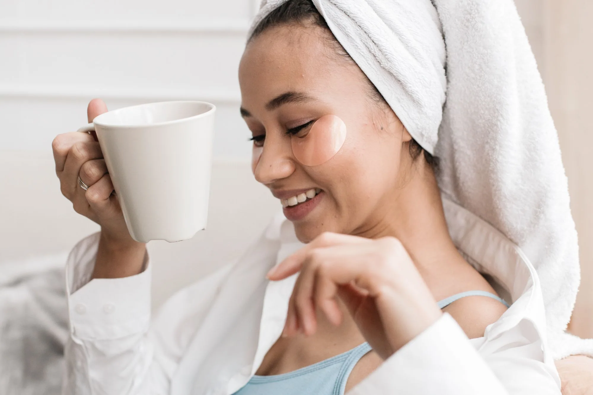 Woman with towel and eye patches enjoying coffee, embracing morning self-care routine. Editorial photograph for our care break guide for Dubai shoppers.