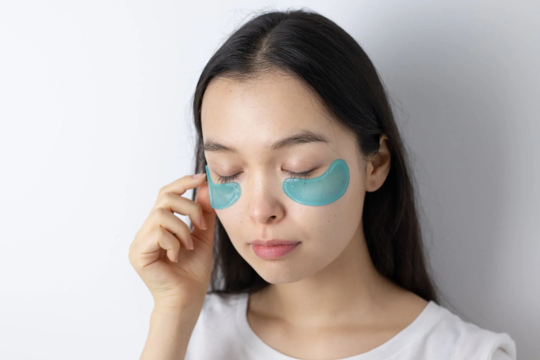 Serene Asian woman with closed eyes applying blue under-eye patches for skincare routine. Editorial illustration for AXXZIA vs Shiseido in Dubai.