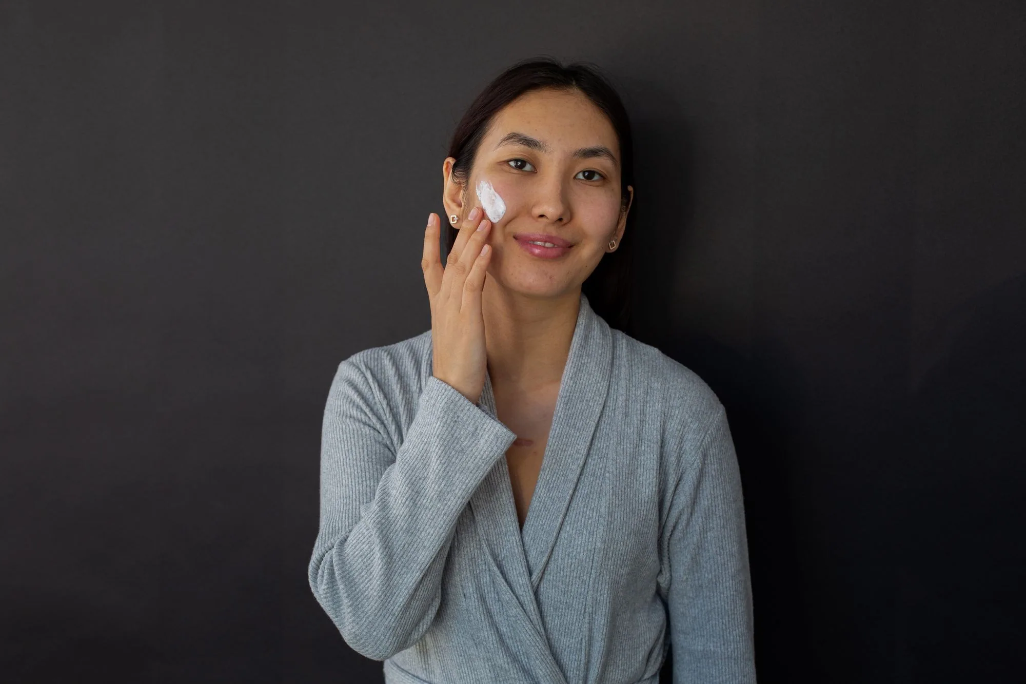 Young Asian woman in bathrobe smiling while applying cream on her face against a black background. Editorial photograph for our Perfect Gel Mask guide for Dubai shoppers.