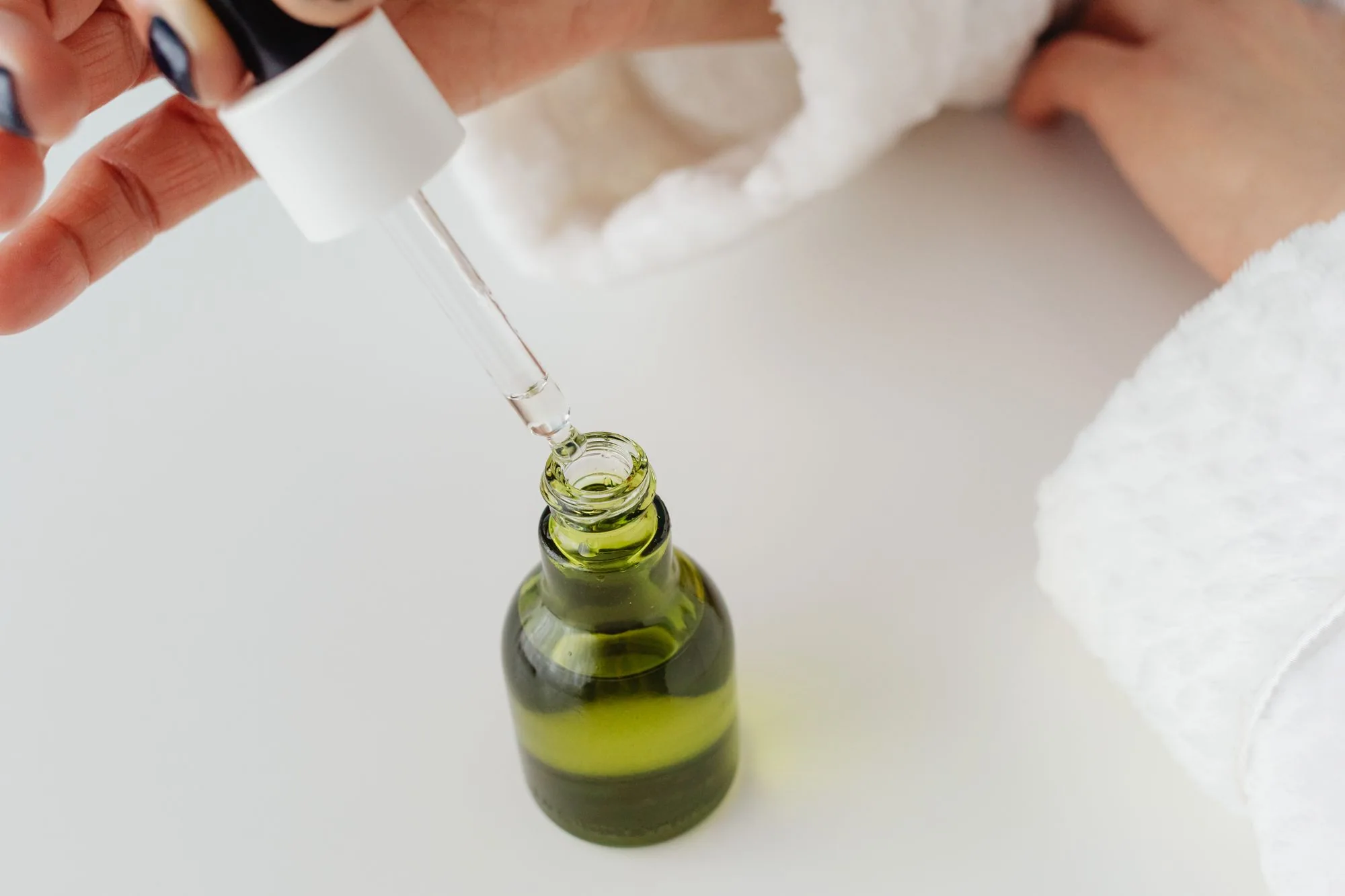 Close-up of hands with manicured nails holding a dropper over a green serum bottle. Editorial photograph for our office skincare guide for Dubai shoppers.