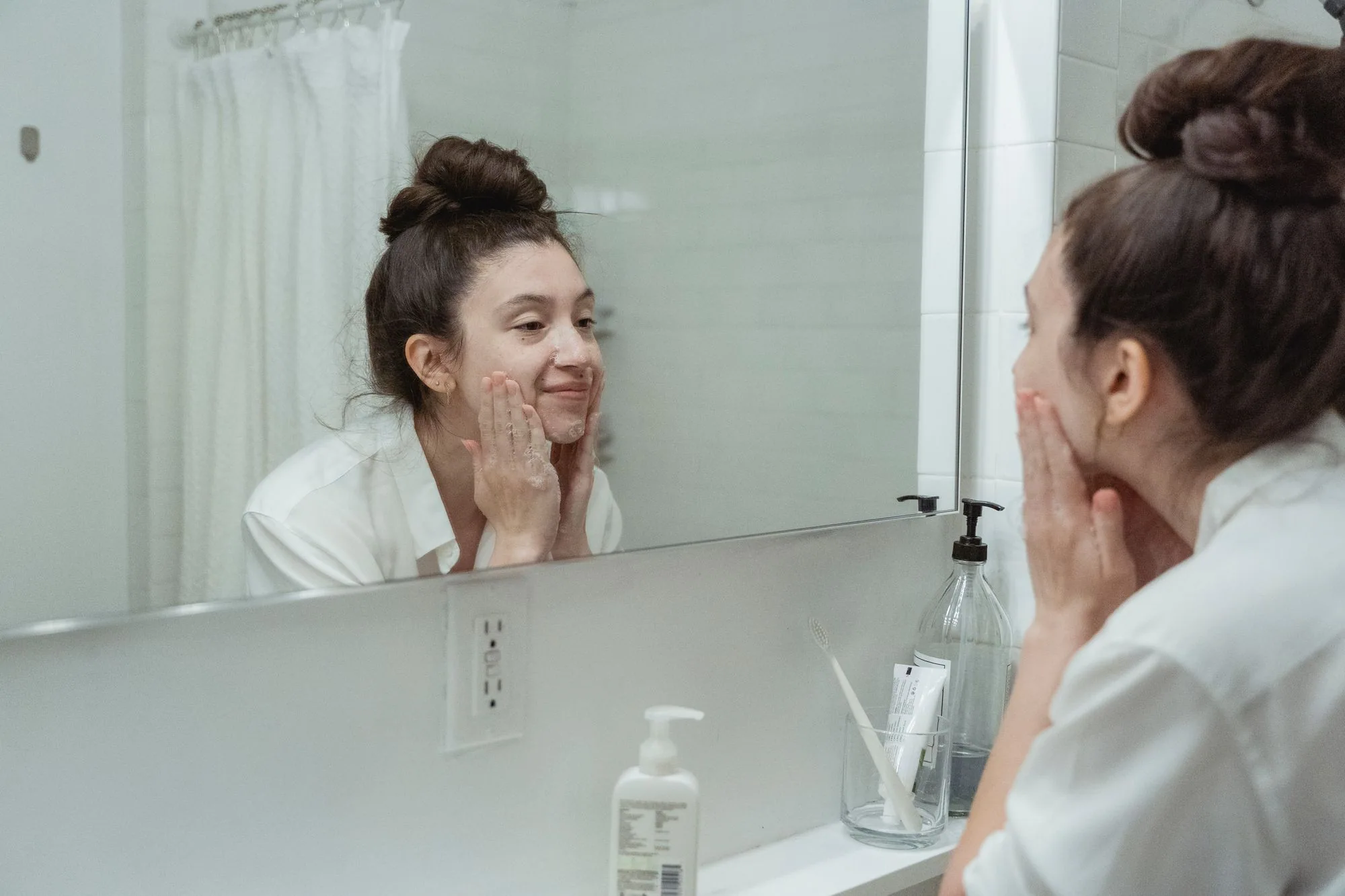 Woman performing skincare routine in a bathroom, applying face cream while looking into a mirror. Editorial photograph for our Clear Wash Foam guide for Dubai shoppers.