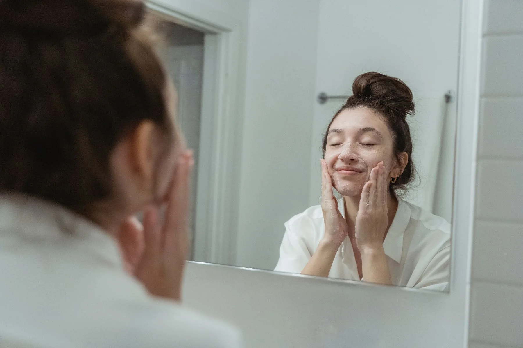Woman applying soap to her face, reflected in the bathroom mirror, enjoying a skincare routine. Editorial illustration for AGTHEORY vs CeraVe vs Shiseido in Dubai.