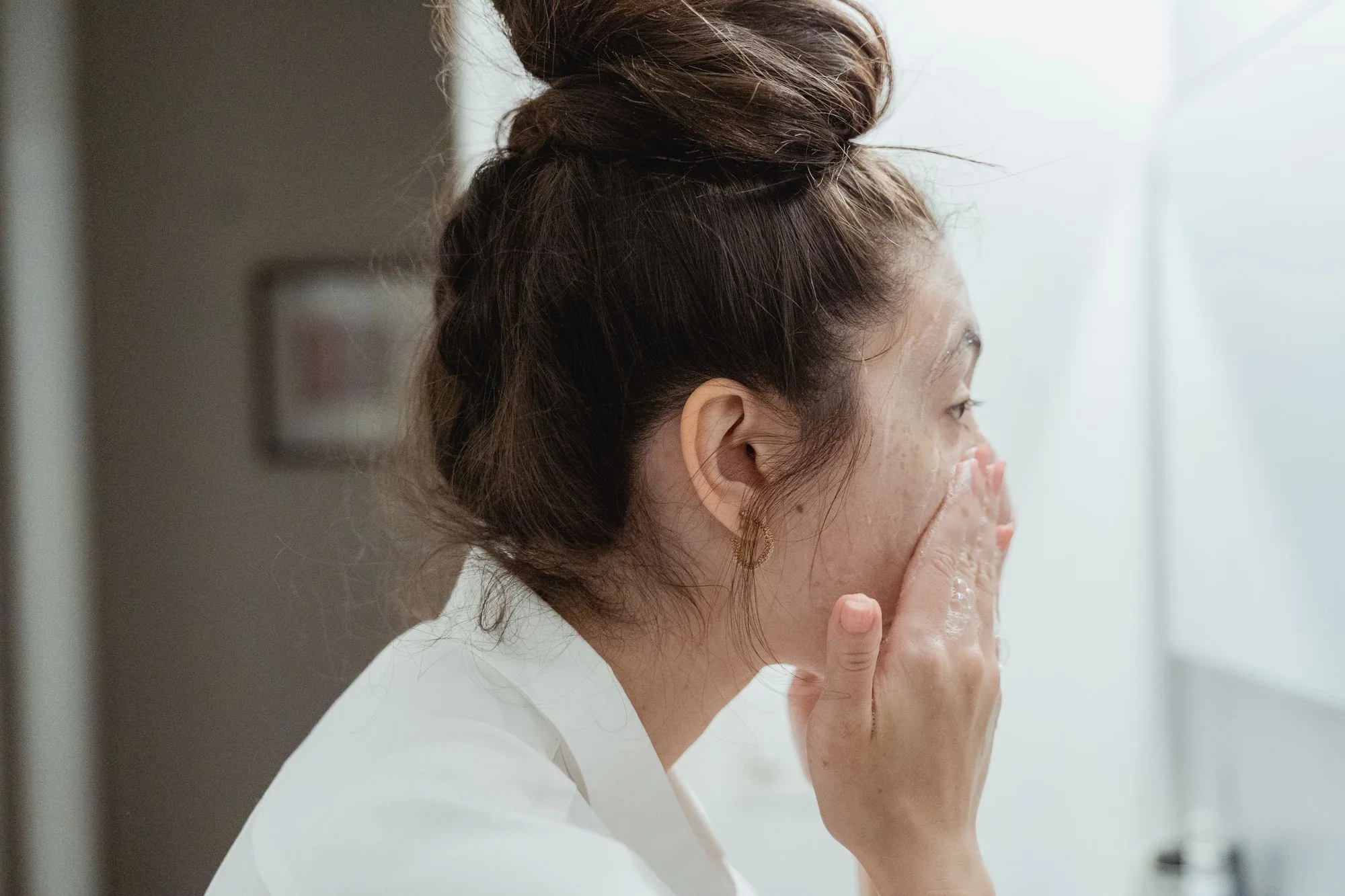 A young woman in a bathrobe follows her skincare routine, washing her face in a bathroom. Editorial photograph for our Deep Cleansing Gel guide for Dubai shoppers.