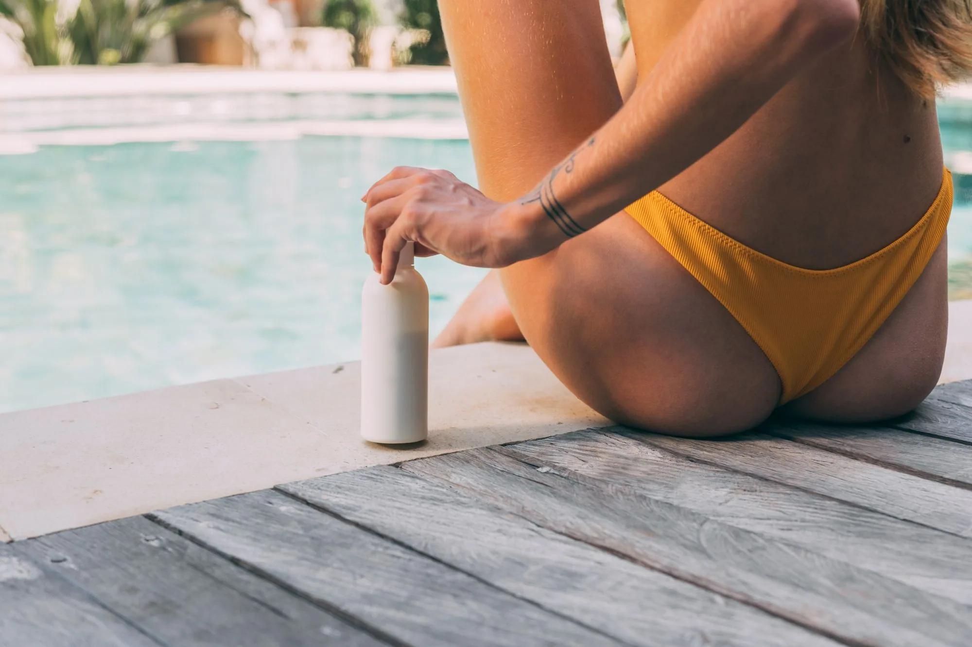 A woman sitting by the poolside, holding a sunscreen bottle in summer sunlight. Editorial photograph for our pool day guide for Dubai shoppers.