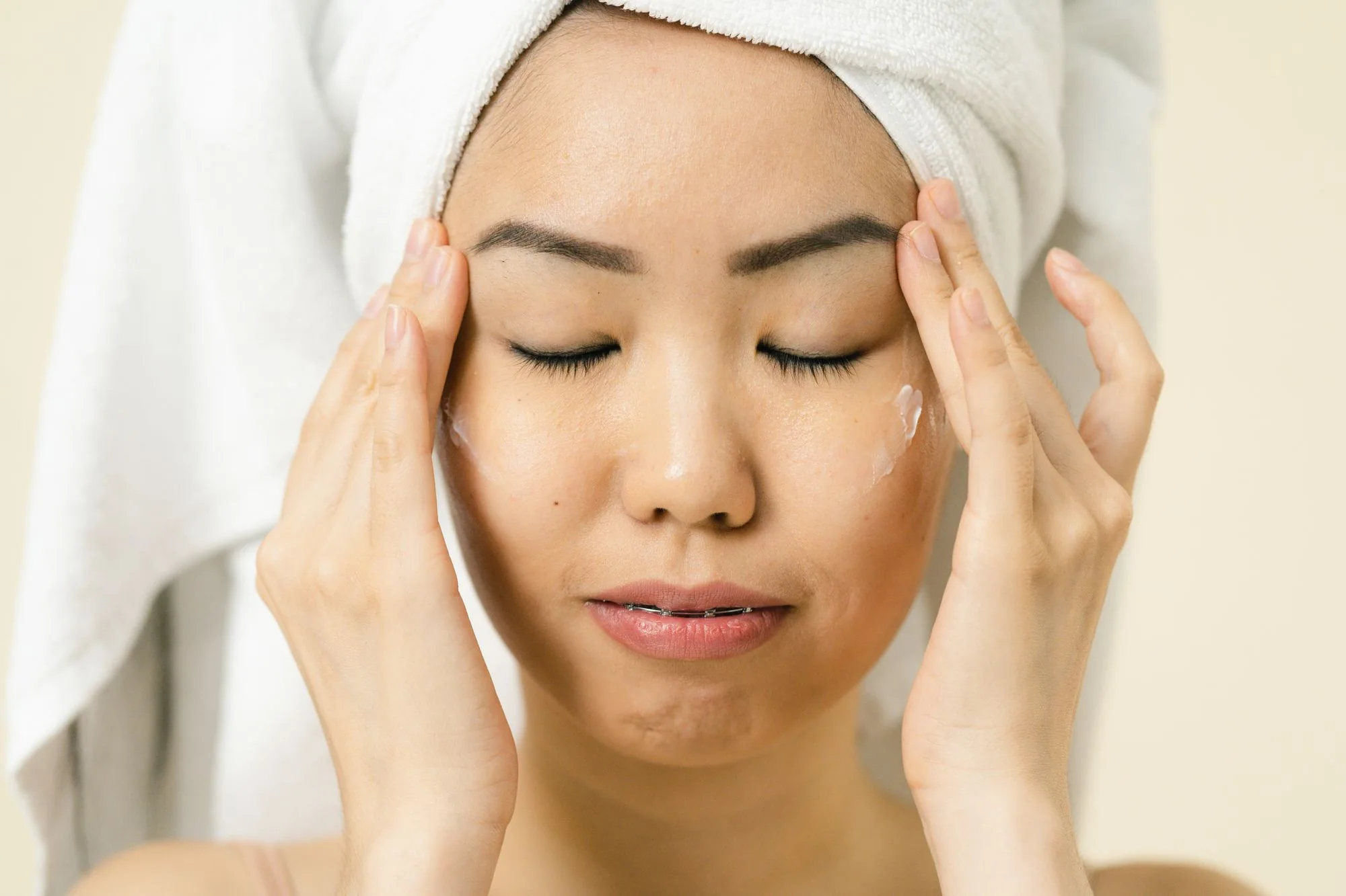 Asian woman with a towel on head applying moisturizer for skincare routine. Editorial photograph for our Silky Emulsion guide for Dubai shoppers.