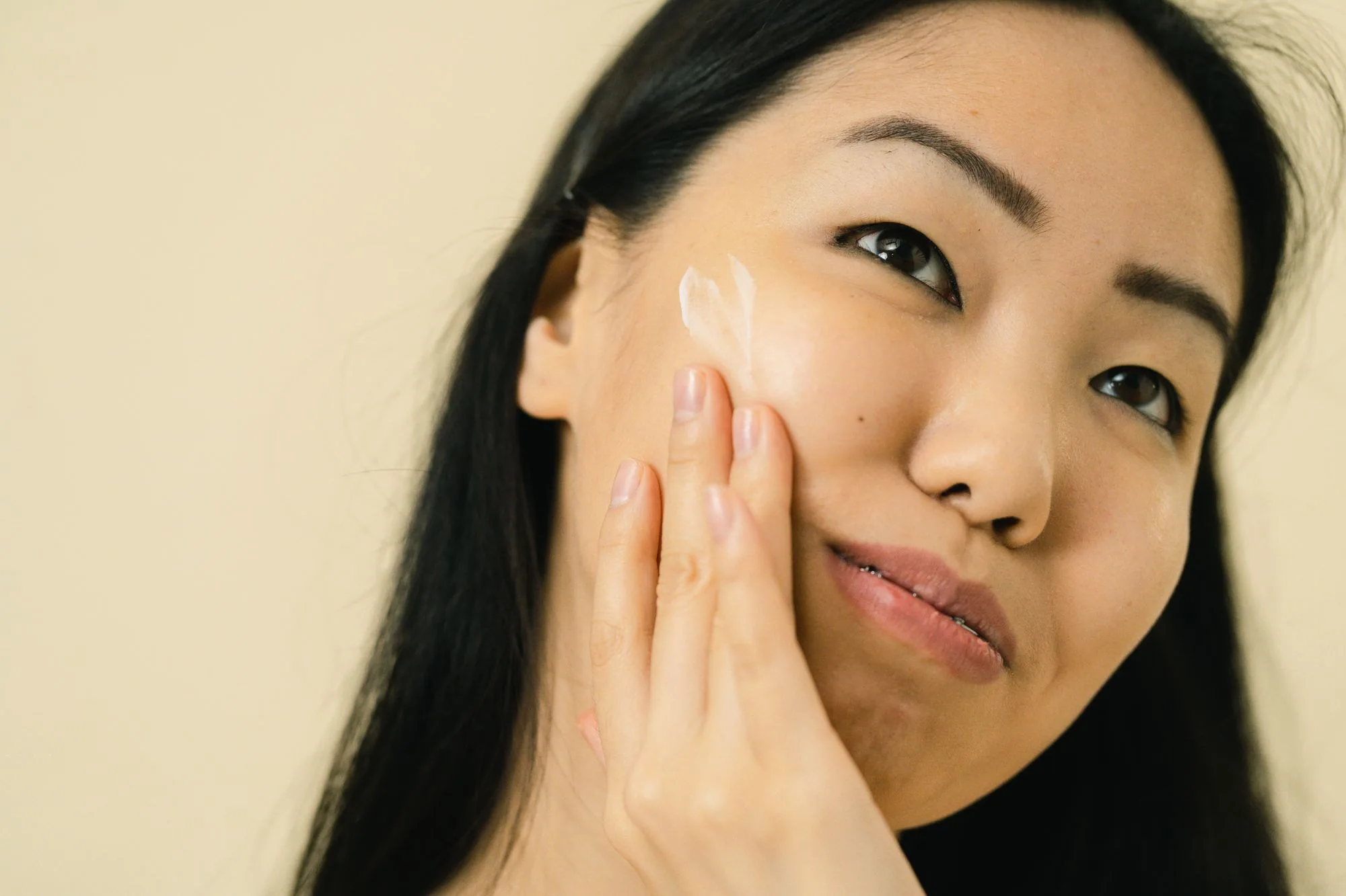 Woman with long black hair applying facial cream in studio. Focus on skincare and beauty routine. Editorial photograph for our one product at a time guide for Dubai shoppers.