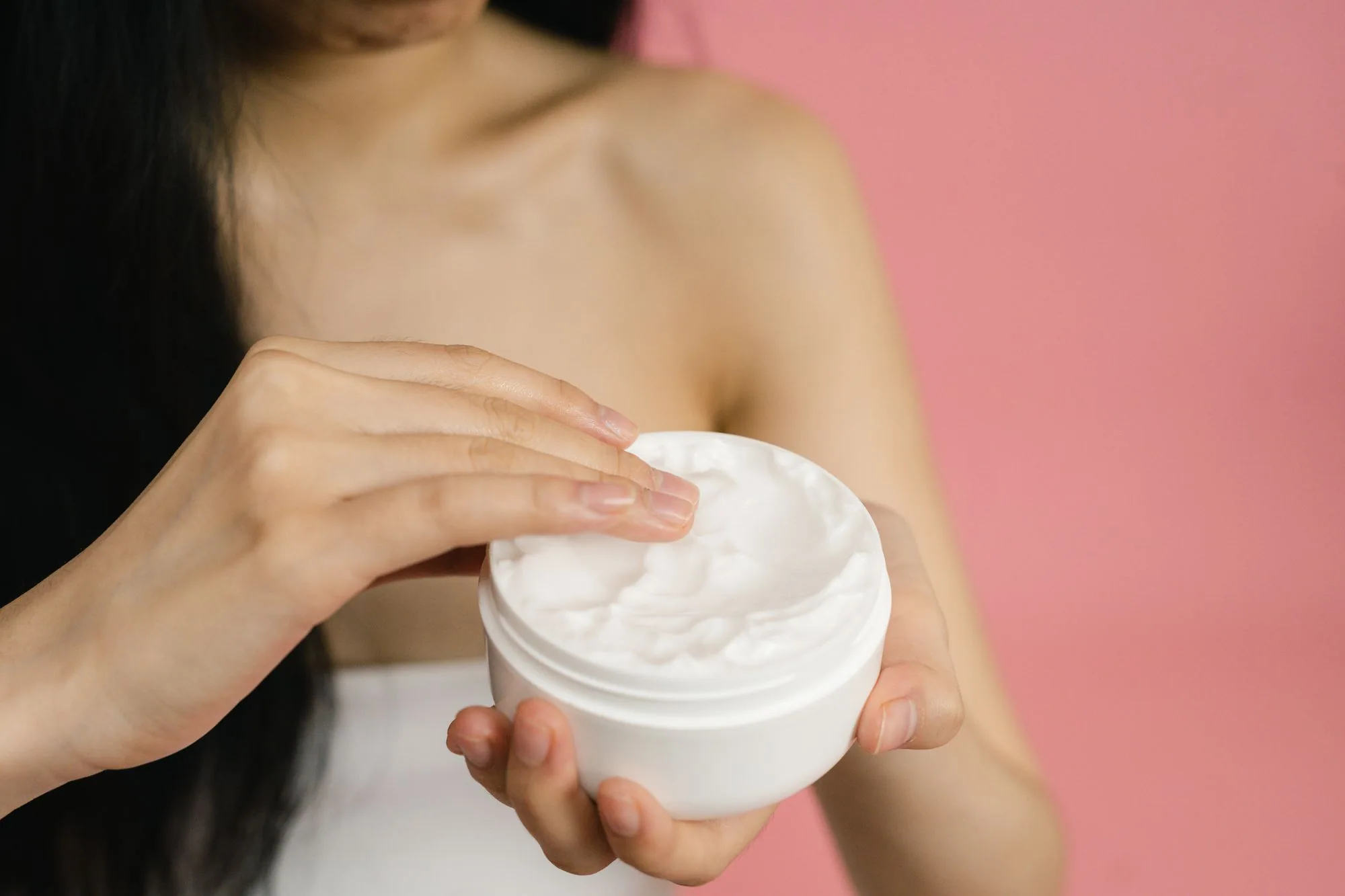 Close-up of a woman applying white cream from a jar against a pink background. Editorial photograph for our emulsion guide for Dubai shoppers.