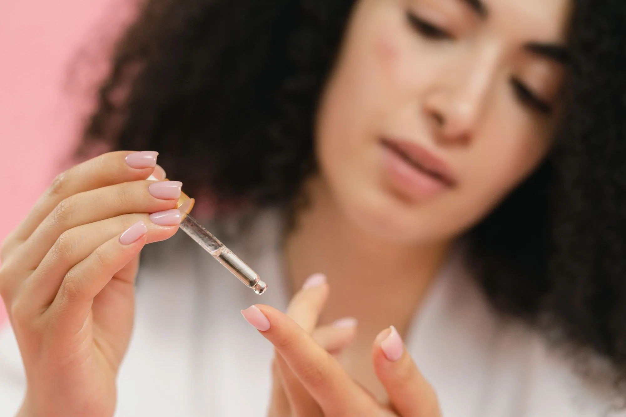 Close-up of a young woman applying skincare serum with a pipette against a pink background. Editorial photograph for our oil essence guide for Dubai shoppers.